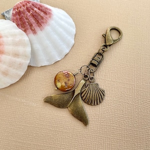 May include: A bronze key chain with a seashell charm, a brown pearl, and a bronze whale tail charm. The key chain is hanging from a white and pink seashell.