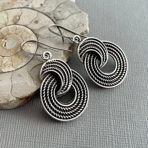 May include: A pair of silver dangle earrings with a twisted, rope-like design. The earrings are circular and have a delicate, intricate pattern.