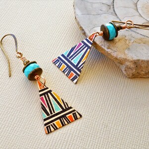 May include: A pair of triangle-shaped earrings with a geometric pattern in black, orange, and turquoise. The earrings are made of metal and have a turquoise bead and a bronze bead.
