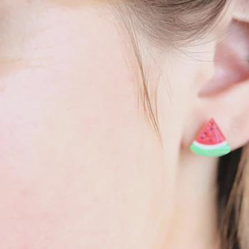 K&ouml;nnte beinhalten: Nahaufnahme eines Ohrs mit einem Ohrstecker in Form einer Wassermelonen-Scheibe. Der Ohrring hat ein rotes Innere mit schwarzen Samen, eine wei&szlig;e Schale und einen gr&uuml;nen Rand. Ein lustiges, sommerliches Accessoire.