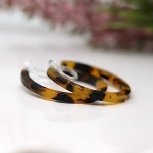 May include: A pair of hoop earrings with a tortoiseshell pattern. The earrings are a warm blend of brown and black, with a clear plastic post. The hoops are a classic, everyday accessory.