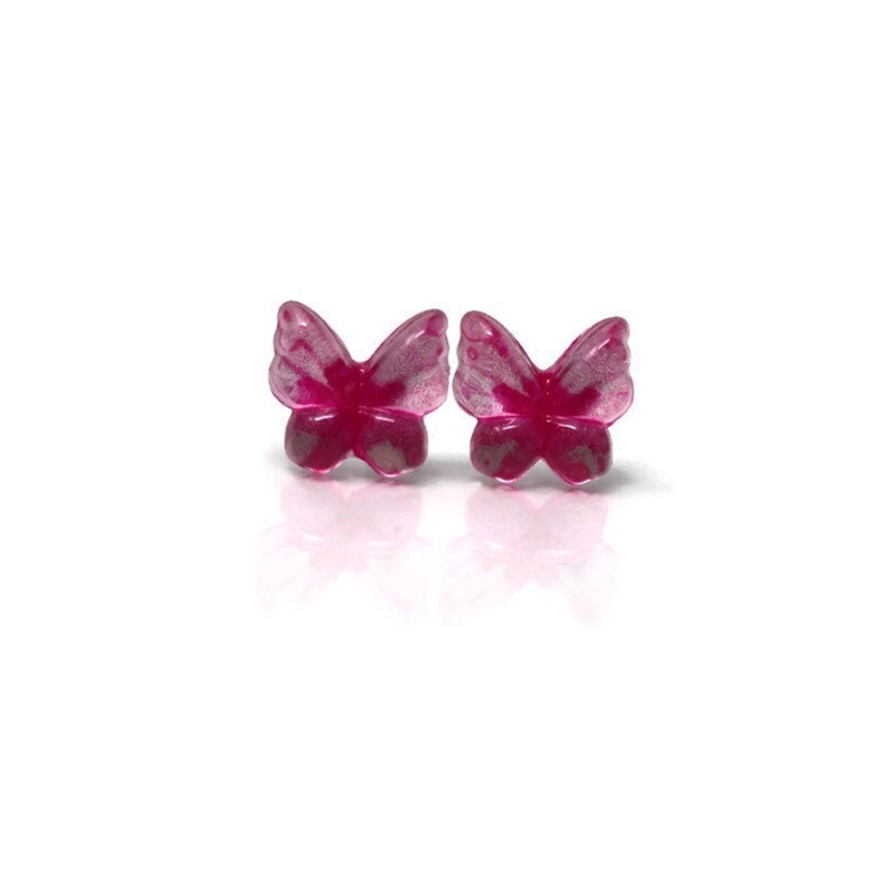 May include: A pair of magenta butterfly-shaped stud earrings. The earrings have a translucent appearance with a hint of silver detailing on the wings. The earrings are set against a white background.