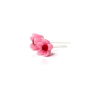 May include: Two small, pink flower-shaped earrings with a dark red center. The earrings are set against a white background, highlighting their delicate design. The petals are slightly translucent, giving them a soft appearance. These earrings are a floral accessory.