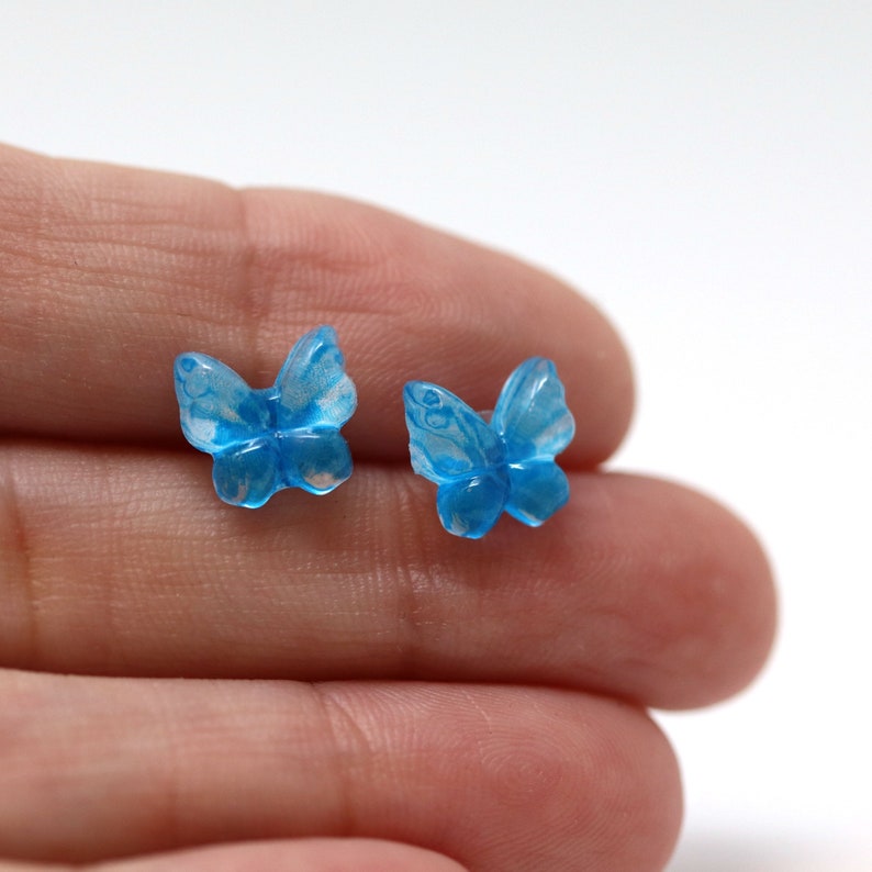 May include: Two small, translucent blue butterfly-shaped earrings. The earrings have detailed wing patterns and are set against a white background. The earrings are small, delicate, and appear to be made of glass or a similar material.