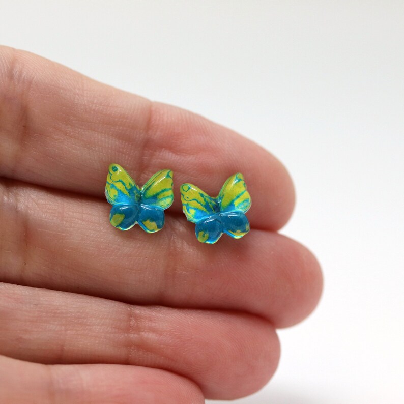 May include: A pair of butterfly-shaped stud earrings. Each earring features a vibrant design with yellow and blue marbling. The earrings are small, delicate, and likely made of resin or a similar material. The butterfly wings are detailed.