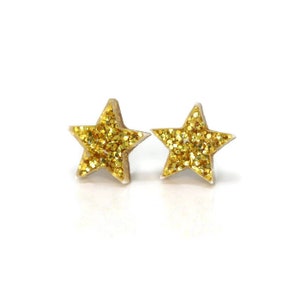 Pu&ograve; includere: Un paio di orecchini a bottone a forma di stella con glitter dorati. Gli orecchini sono realizzati in un materiale trasparente con glitter dorati che riempiono la forma a stella. Su sfondo bianco.