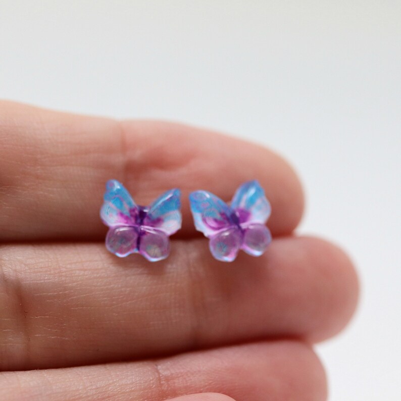 May include: Two small butterfly-shaped accessories. The wings are a translucent blue with purple accents, and the bodies are a translucent purple. The butterflies are small and delicate, with a whimsical design.