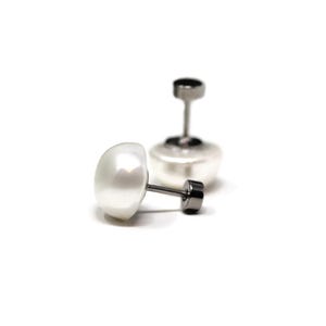 May include: Two white pearl stud earrings with dark gray posts and backs. One earring is lying on its side, while the other is upright in the background. The earrings have a classic, elegant design.
