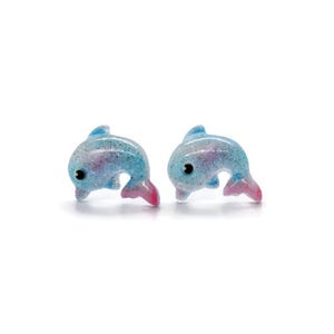May include: A pair of dolphin-shaped earrings. Each earring is a light blue color with glitter and pink accents on the tail. The dolphins have black eyes and are set against a white background.