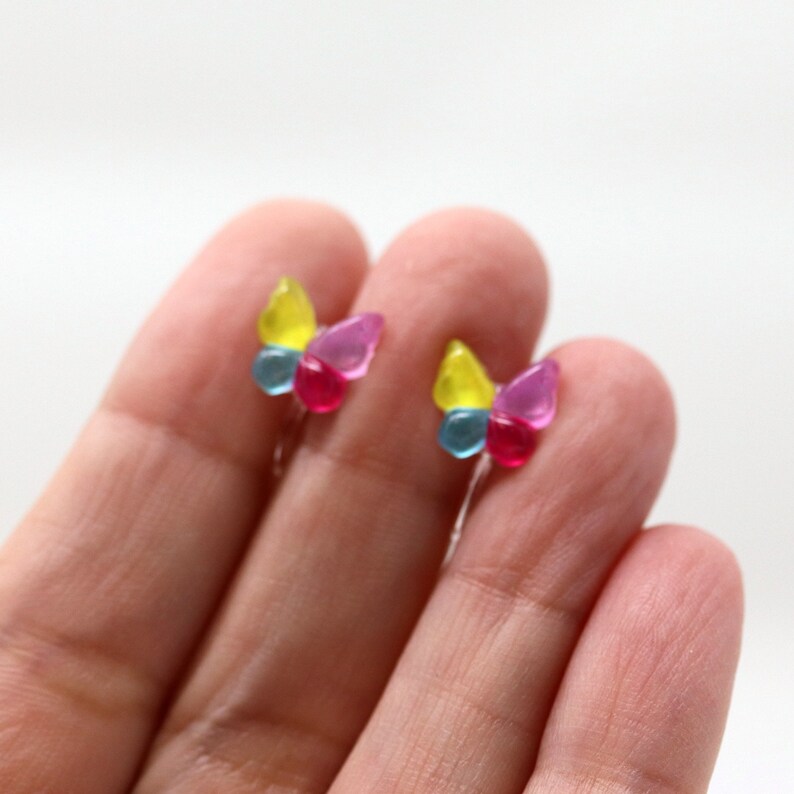 May include: A pair of colorful butterfly earrings. Each earring features a yellow, blue, and pink wing design. The earrings are held by a person's fingers against a white background.