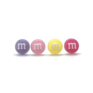 May include: Four colorful, round candies in a row. The candies are purple, pink, yellow, and red. Each candy has a white "m" printed on it. The candies have a glittery texture.