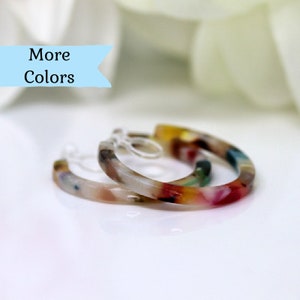 May include: A pair of colorful hoop earrings with a marbled design. The earrings feature a mix of colors including red, yellow, green, and blue. The text "More Colors" is displayed on a light blue banner.