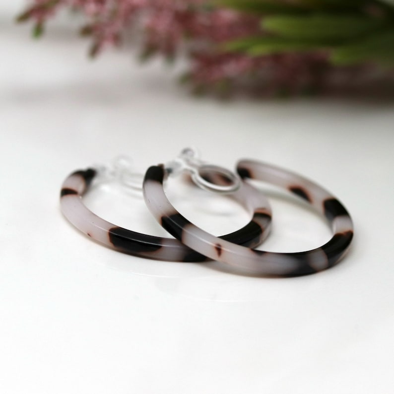 May include: A pair of hoop earrings with a marbled design. The earrings feature a combination of black, white, and light brown colors. The earrings are made of a lightweight material and have a clip-on closure.