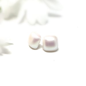 May include: Two white, square-shaped beads with a pearlescent sheen. The beads are slightly rounded and sit on a white background. The beads have a subtle pink hue and are likely used for jewelry making or crafting.