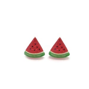Peut inclure: Deux boucles d'oreilles en forme de tranches de pastèque. Chaque boucle d'oreille est un triangle rouge avec des détails de graines noires et une peau verte. Sur fond blanc.