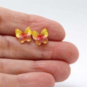 May include: Two small, translucent butterfly-shaped beads. The beads are yellow with red and yellow accents. The beads are resting on a light-colored surface.