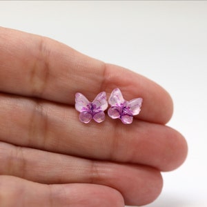 May include: Two small, translucent butterfly-shaped embellishments. The wings are a pale pink with darker purple veining and a purple center. The butterflies are held in a hand against a white background.