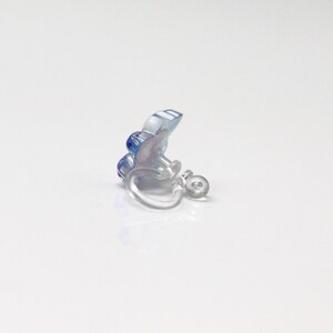 May include: A clear clip-on earring with a blue and purple floral design. The earring has a small, round, clear clip mechanism. The flower has a translucent appearance, with visible details.