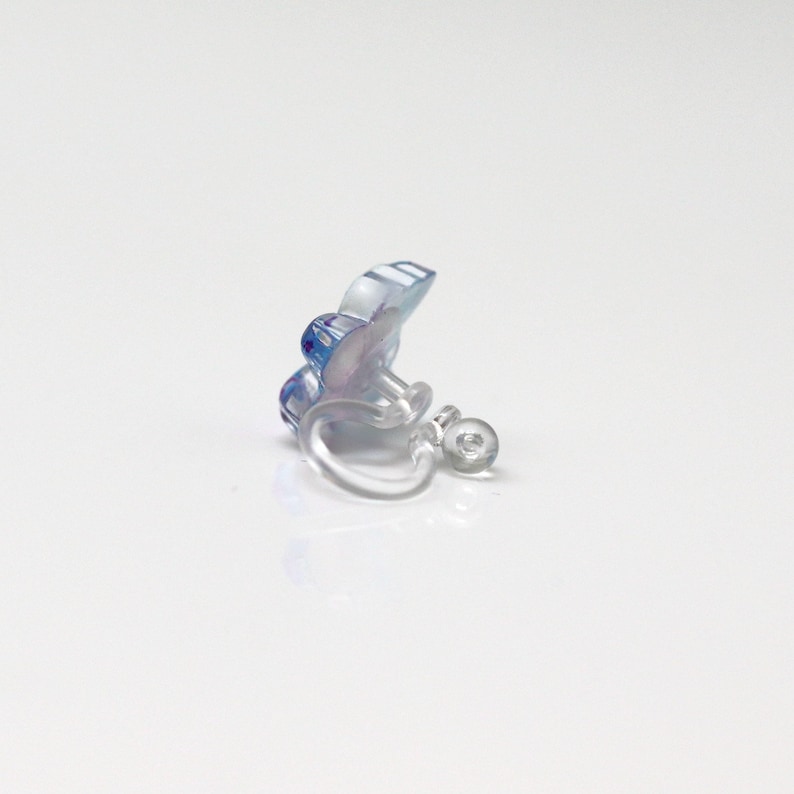 May include: A clear clip-on earring with a blue and purple floral design. The earring has a small, round, clear clip mechanism. The flower has a translucent appearance, with visible details.