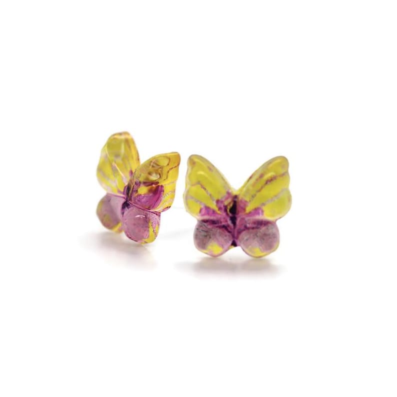 May include: A pair of butterfly-shaped stud earrings. The earrings feature yellow wings with purple accents and a purple body. The earrings are set against a white background.