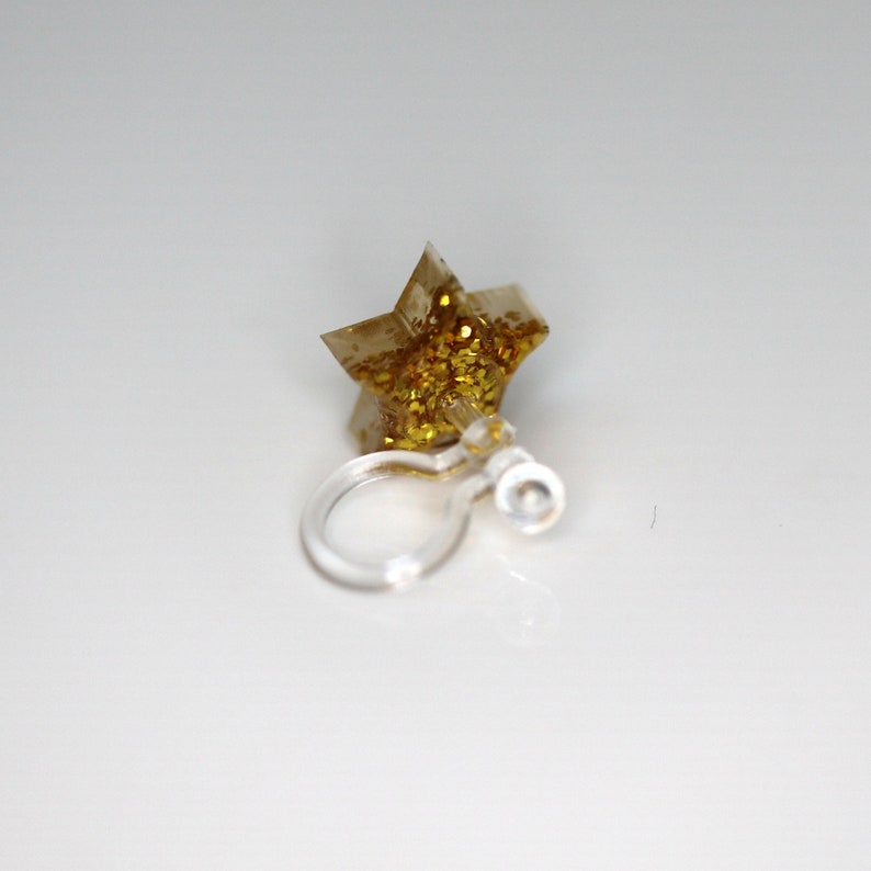 Pu&ograve; includere: Orecchino a clip trasparente con un ciondolo a forma di stella. La stella &egrave; traslucida con glitter dorati incorporati. La clip dell'orecchino &egrave; in plastica trasparente.