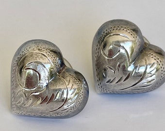 May include: Two silver heart-shaped earrings. Each earring has an engraved floral design and a dotted border. The earrings are displayed on a white surface, and the metal has a polished finish.