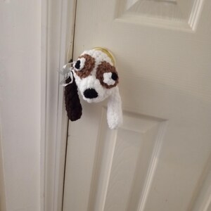 May include: A handmade crochet dog head key holder in white, brown, and black, hanging on a door. The dog has large brown eyes and a black nose. The key holder is attached to a set of keys.