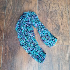 May include: A hand-knitted scarf in shades of teal, purple, and green. The chunky knit scarf is made of a soft, textured yarn. The scarf is lying on a wooden surface.