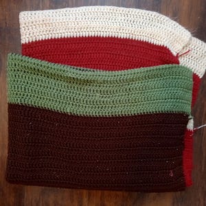 Può includere: Articoli all'uncinetto fatti a mano in crema, rosso, verde e marrone. Gli articoli sembrano essere una sciarpa o un collo, con un design a righe. Il lavoro all'uncinetto è visibile, mostrando la trama e il motivo dei punti. Gli articoli sono piegati e impilati.
