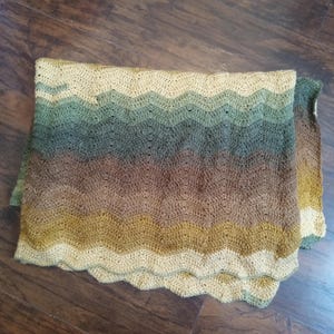 May include: A folded, handmade crochet blanket with a wavy pattern. The blanket features a gradient of colours, transitioning from light yellow to green, then brown, and back to yellow. The edges have a scalloped design.