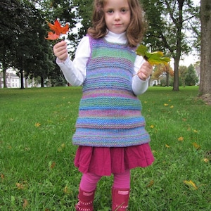 May include: A young girl wearing a striped knitted vest and a pink skirt holds autumn leaves in a grassy field.