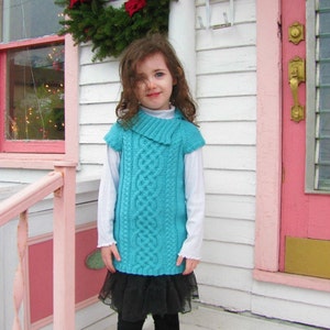 May include: A young girl wearing a teal blue knitted sweater vest with a cable knit pattern. She is wearing a white long-sleeved shirt underneath and a black tulle skirt. The girl is standing in front of a pink door with a wreath on the wall behind her.