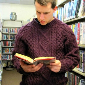 Può includere: Un uomo con un maglione di lana a trecce viola sta leggendo un libro in una biblioteca. È in piedi di fronte a una libreria piena di libri.