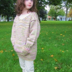May include: A young girl wearing a multi-colored striped knitted sweater with a v-neck and a pocket on each side. She is wearing white pants and standing in a grassy field.