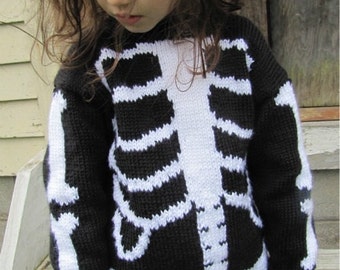 Könnte beinhalten: Ein junges Mädchen trägt einen schwarz-weißen Strickpullover mit einem Skelett-Design. Der Pullover hat lange Ärmel und einen Rundhalsausschnitt. Das Mädchen trägt eine blaue Jeans.