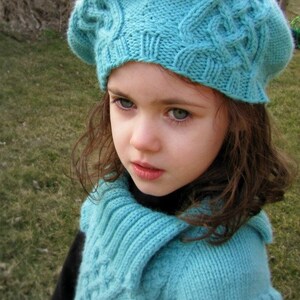 May include: A young girl wearing a light blue knitted hat and matching sweater. The hat and sweater have a cable knit pattern.