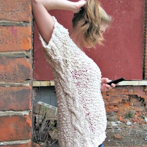 May include: A woman wearing a white knitted sweater with a cable knit pattern and blue jeans. She is standing in front of a brick wall and holding a cell phone.