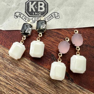 May include: Close-up of two pairs of earrings. One pair features a black and clear top stone with a white, faceted, rectangular bottom stone. The other pair has a pink oval top stone and a white, faceted, rectangular bottom stone. Gold-colored metal findings.