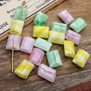 May include: A selection of colourful, rectangular glass beads in shades of yellow, green, and pink. Some beads have a swirled pattern. A gold pin is inserted through a few of the beads. The beads are scattered on a wooden surface.