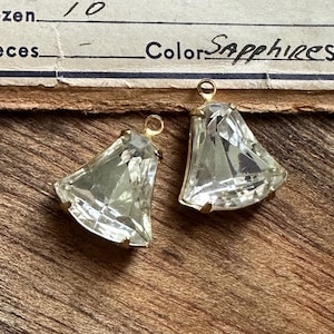 Pode incluir: Dois pingentes de pedras preciosas claras e facetadas com engastes dourados. As pedras preciosas têm formato de sino com uma argola no topo para pendurar. A palavra "Sapphires" está escrita em um pedaço de papel no fundo.