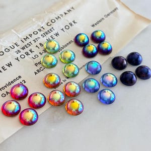 May include: A collection of round, iridescent cabochon buttons in shades of red, orange, yellow, green, blue, and purple, arranged on a vintage paper envelope labeled VOGUE IMPORT COMPANY 28 WEST 37th STREET NEW YORK 18 Western Germany.