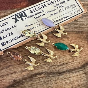 May include: Close-up of several vintage jewelry components. Each piece features a gold-colored bird charm connected to a gemstone in various colors, including green, yellow, and purple. The text on the vintage tag reads "GEORGE MELLON" and "Sapphire Size 18 MM".