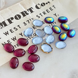 May include: A collection of oval cabochons in various colors including translucent light blue, deep magenta, and iridescent purple-green. One light blue cabochon is set in a gold-toned metal bezel. The items are arranged on a vintage paper label.