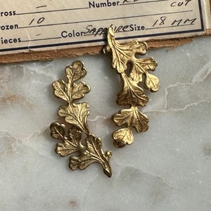May include: Two gold-colored leaf-shaped ornaments. The leaves have detailed veining and are arranged in a curved, branch-like design. The size is 18 mm. The background is a light-colored, marbled surface.