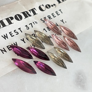 May include: A collection of marquise-cut faceted crystals in shades of deep magenta, light pink, and smoky grey. The crystals are arranged on a white paper background featuring printed text that reads IMPORT CO., WEST 37th STREET, NEW YORK, N.Y.