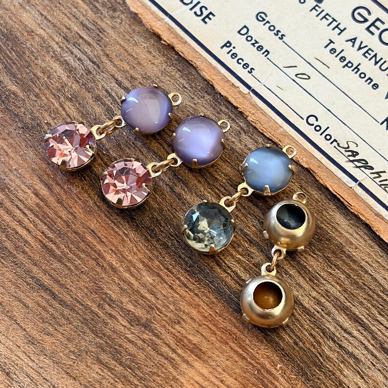 May include: Close-up of several pairs of vintage earrings with gold-tone metal settings. The earrings feature a variety of colourful gemstones, including pink, purple, blue, and clear stones. The earrings are displayed on a wooden surface.
