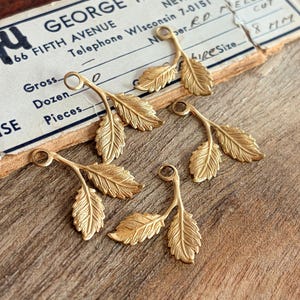 May include: Five gold-toned metal charms, each shaped like a small branch with two textured leaves and a circular loop at the top for attachment. The charms are arranged on a vintage paper label with printed text including George and Fifth Avenue.