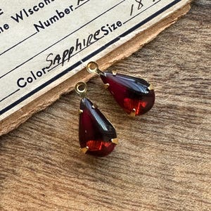 May include: Two teardrop-shaped earrings with a deep red gemstone and gold-colored metal settings. The earrings are displayed on a textured, brown surface, with a vintage document visible in the background. The document has the word "Sapphire" written on it.