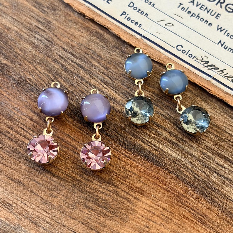 May include: Four pairs of dangling earrings with gold-coloured metal findings are displayed. Each earring has a top stone in grey or purple, connected to a lower, faceted stone in pink or green. The earrings are set on a wooden surface.