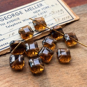May include: A collection of translucent amber-colored glass beads with a square, four-lobed shape. Some beads are strung on a thin gold-toned wire, while others are loose. The beads are displayed on a vintage paper label marked GEORGE MELLER.